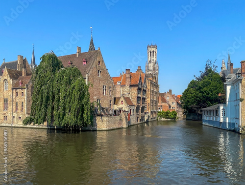 Brugge, Flanders, Belgium - June 25, 2024: Halletoren over historic buildings and Dijver canal under blue sky seen from Rozenhoedkaai, photo