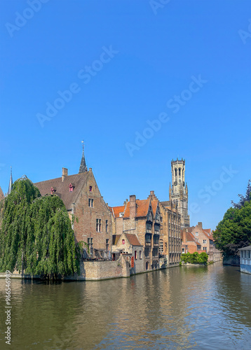 Brugge, Flanders, Belgium - June 25, 2024: Halletoren over historic buildings and Dijver canal under blue sky seen from Rozenhoedkaai, photo