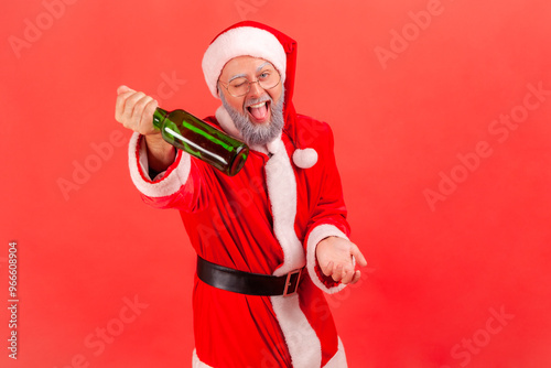 Happy funny elderly man with gray beard wearing santa claus costume standing with alcohol bottle and offering beer, celebrating new year. Indoor studio shot isolated on red background. photo