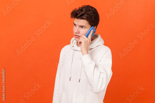 Portrait of serious handsome young man wearing white hoodie standing with smartphone in hands, making call and talking with friend. Indoor studio shot isolated on orange background.