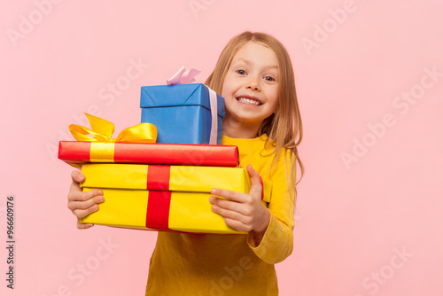 Portrait of joyful cheerful blonde little girl celebrating her birthday holding stack of presents, smiling, wearing yellow jumper. Indoor studio shot isolated on pink background.