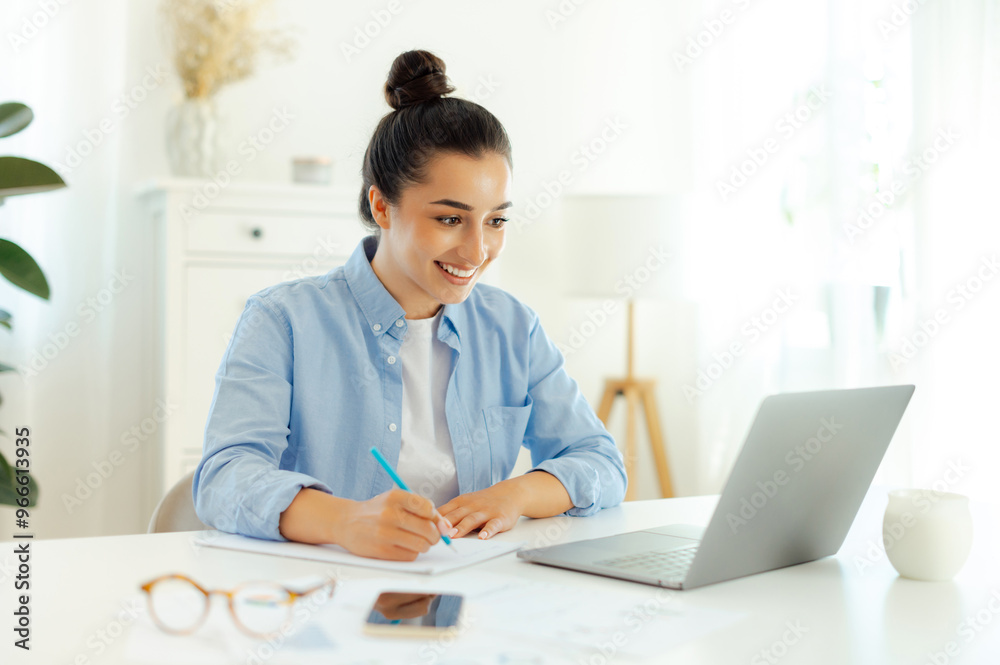Online working. Successful smiling arabian or indian woman in a blue shirt, sits a table, working on the laptop, develop new digital project, planning marketing strategy, takes notes in a notebook Online working. Successful smiling arabian or indian woman in a blue shirt, sits a table, working on the laptop, develop new digital project, planning marketing strategy, takes notes in a notebook