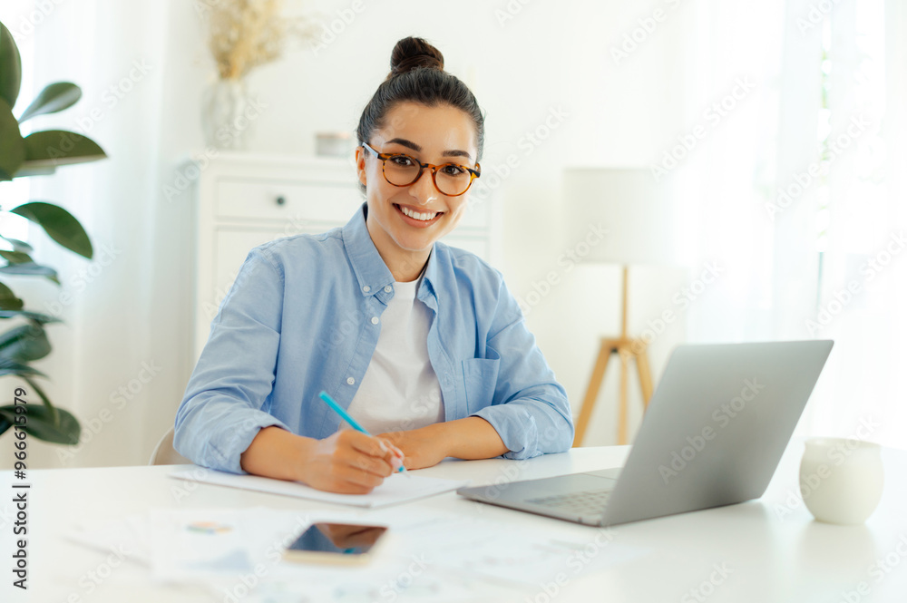 Photo of a confident pretty woman. Successful arabian or indian woman in a blue shirt, working on the laptop, develop new digital project, planning marketing strategy, looks and smiles at camera Photo of a confident pretty woman. Successful arabian or indian woman in a blue shirt, working on the laptop, develop new digital project, planning marketing strategy, looks and smiles at camera