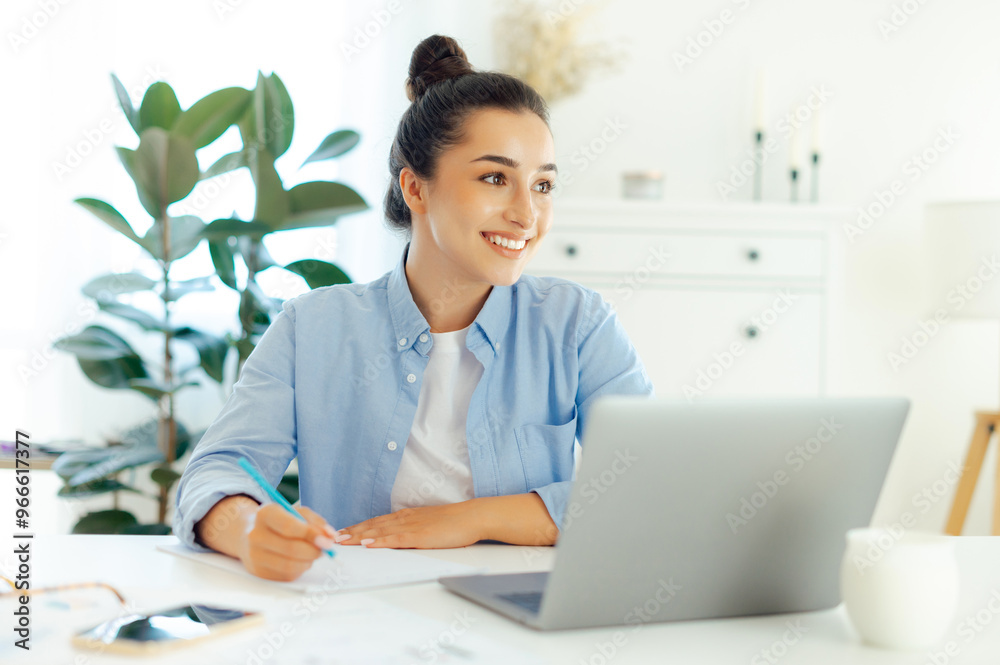 Positive charming confident arabian or indian business woman, project manager, company ceo, sits at a desk, working on a laptop, planning strategy and business plan, takes notes, looks away and smiles Positive charming confident arabian or indian business woman, project manager, company ceo, sits at a desk, working on a laptop, planning strategy and business plan, takes notes, looks away and smiles