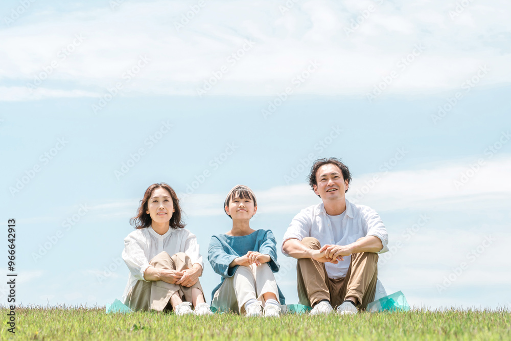 青空の見える公園・丘に座る家族・親子（父親・母親・娘）
