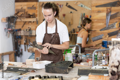 Concentrated young guy in apron viewing interestedly taller mold in workshop photo