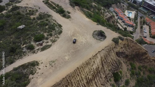 Aerial tracking by drone of a compact blue car, rolling on the ridge of a mountain, with arid vegetation and beach in the background