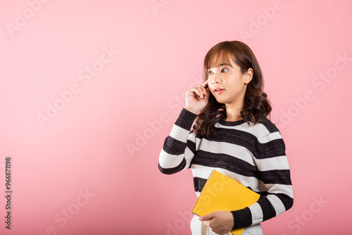 Portrait of beautiful Asian young woman teenage smiling holding a yellow book and reading, studio shot isolated on pink background with copy space