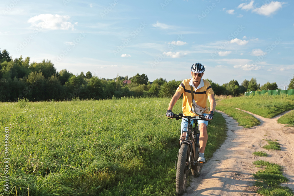 Cyclist in helmet with equipment on electric bicycle rides along path in field in countryside. Sports and health. Summer hobby. Cyclist in helmet with equipment on electric bicycle rides along path in field in countryside. Sports and health. Summer hobby.