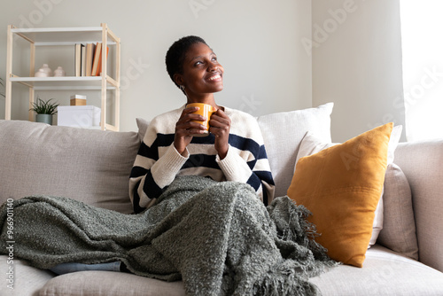 Pensive happy African American young woman relaxing at home, sitting on the sofa drinking coffee looking out the window