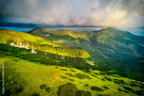 Breathtaking aerial view of green, rugged mountain peaks under dramatic sky. Scene captures contrast between vibrant landscape and moody clouds, with sunlight breaking through. Carpathians, Chornogora