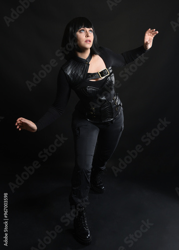 Full length portrait of beautiful black female model with short black hair wearing sci fi futuristic dystopian leather bodysuit costume. Standing pose, isolated dark studio background moody silhouette