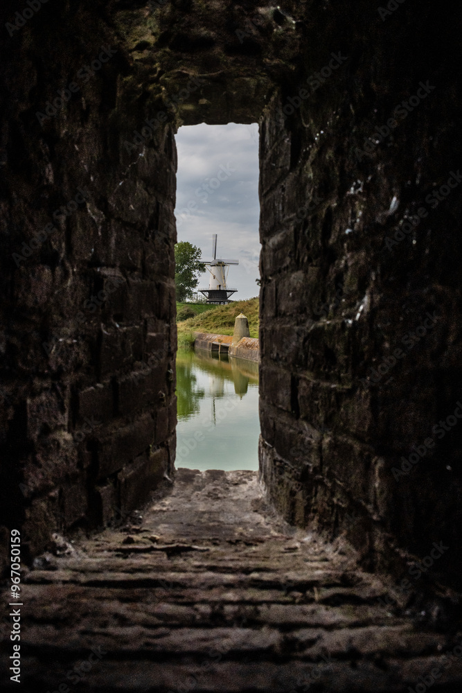 De Koe windmill viewed through window in wall of Stenen Beer stone bear fortified passageway walkable tunnel. historic landmark building present functional Dutch architecture De Koe windmill viewed through window in wall of Stenen Beer stone bear fortified passageway walkable tunnel. historic landmark building present functional Dutch architecture