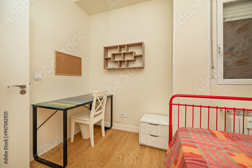 A student bedroom with black metal desk, white wooden chair and red metal bed