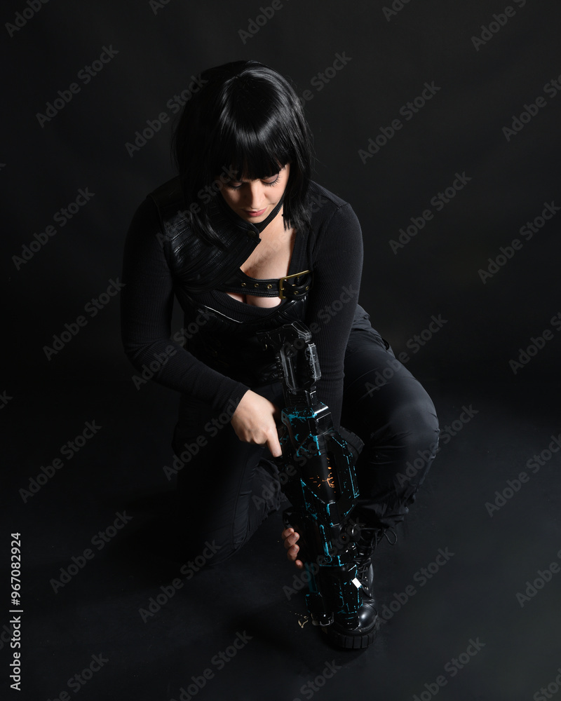 Naklejka premium Full length portrait of beautiful black female model wearing black sci fi futuristic dystopian leather bodysuit costume. Kneeling pose, holding gun weapon. isolated dark studio background moody silhou