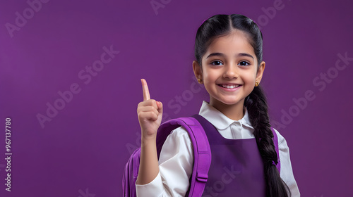 Happy Indian primary school girl in uniform with backpack pointing at copy space for advertising products or services for pupils, isolated on violet background.