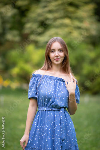 Young smiling girl walks through spring park in good mood. Beautiful girl with long light brown hair in blue dress poses among plants, trees and flowers. Summer fashion, lightness and relaxation.