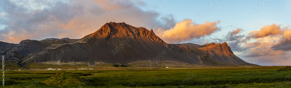 Panoramic photograph of an Icelandic mountain during the golden hour, with orange and pink clouds in the sky and a green meadow in the foreground. Panoramic photograph of an Icelandic mountain during the golden hour, with orange and pink clouds in the sky and a green meadow in the foreground.
