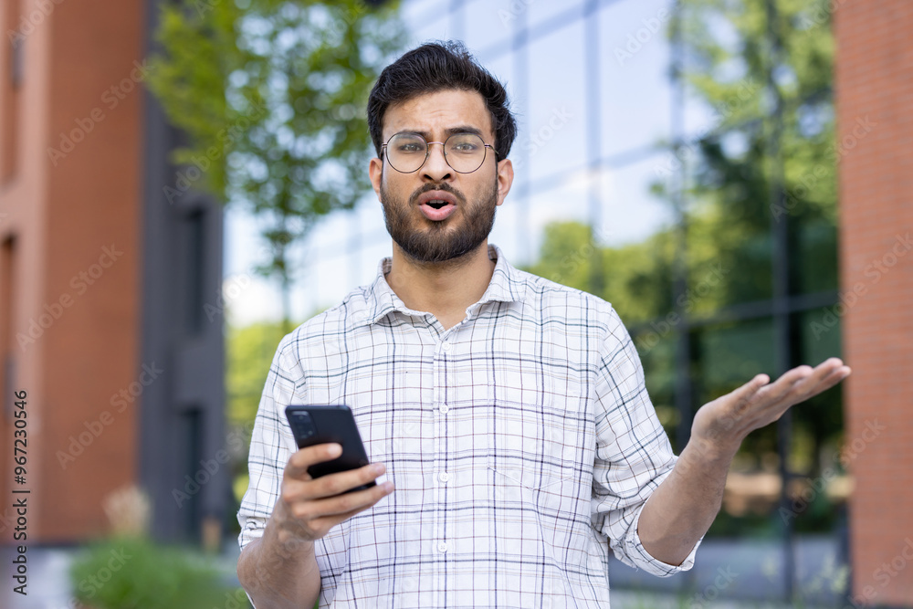 Portrait of frustrated and upset young Muslim man in shirt and bib standing outside office, holding phone, looking at camera and spreading arms Portrait of frustrated and upset young Muslim man in shirt and bib standing outside office, holding phone, looking at camera and spreading arms