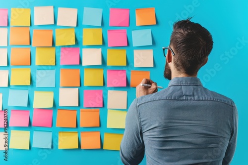 Rear view of a man standing in front of a board covered with colorful sticky notes, brainstorming ideas in a creative workspace or office.