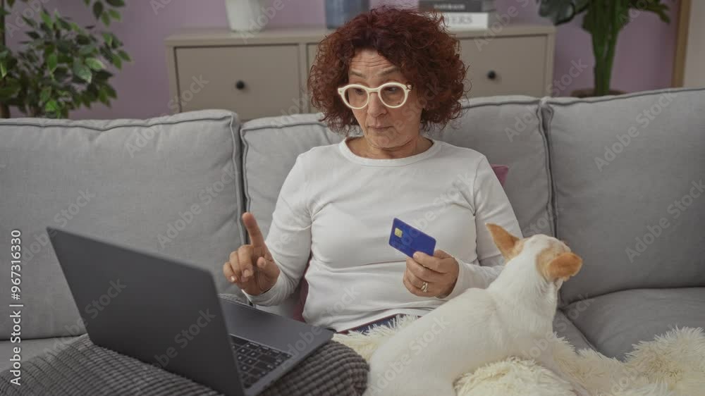 Elderly woman with curly hair and glasses sitting on a couch in a cozy living room, holding a credit card, while using a laptop with a small chihuahua dog beside her. Elderly woman with curly hair and glasses sitting on a couch in a cozy living room, holding a credit card, while using a laptop with a small chihuahua dog beside her.
