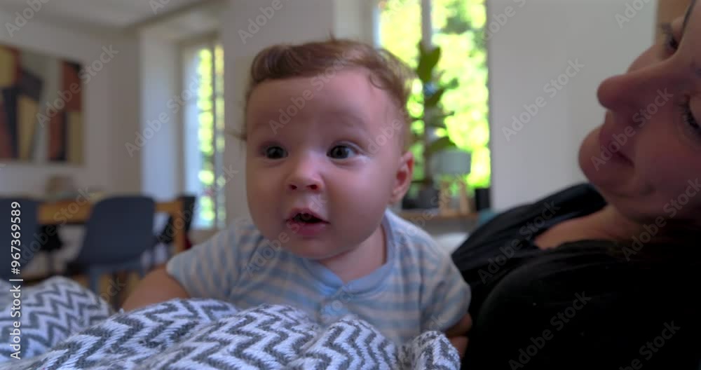 Baby lying on a blanket, looking up with curiosity, while mother smiles in the background in a cozy living room filled with natural light and a warm family atmosphere Baby lying on a blanket, looking up with curiosity, while mother smiles in the background in a cozy living room filled with natural light and a warm family atmosphere