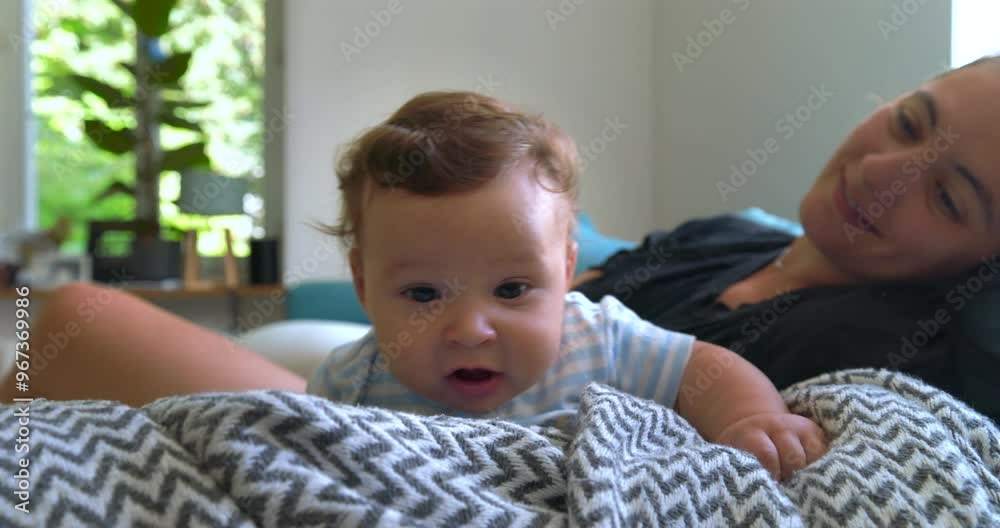 Baby lying on a blanket, exploring and playing while mother watches in the background, warm family moment in a bright and cozy living room, baby's curiosity