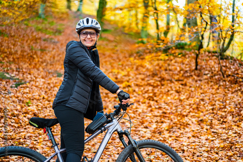  Portrait of beautiful middle aged smiling woman wearing black sporty jacket, black cycling pants and white bike helmet and sunglasses standing with bicycle in forest in autumn scenery.