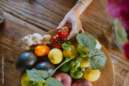 A hand reaches for vibrant fresh produce in a wooden bowl, including tomatoes, lemons, and avocados. photo