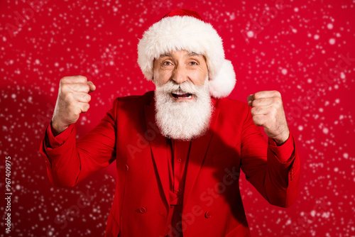 Photo of aggressive angry mature man dressed christmas costume hat rising fists isolated red color background