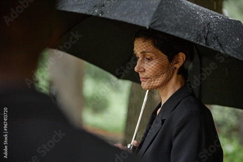 Side view of senior woman in black suit and veil grieving over loss of loved one at memorial ceremony hiding from rain under umbrella at graveyard, copy space
