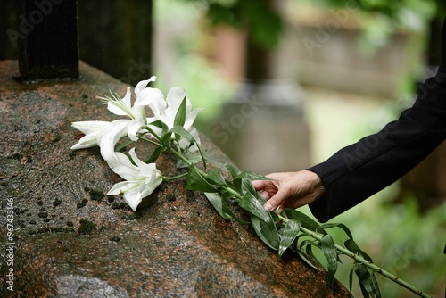 Close up on hand of senior woman placing white lilies on rain wet stone of memorial monument at grave in cemetery commemorating departed loved one, copy space