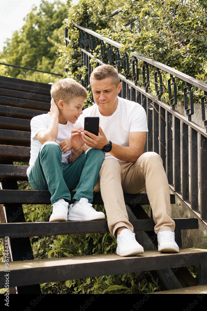 Father and son sitting on steps in a park looking at a smartphone. Father and son ordering, talking online, playing on smartphone. The boy is looking at his father's phone, enjoying their time Father and son sitting on steps in a park looking at a smartphone. Father and son ordering, talking online, playing on smartphone. The boy is looking at his father's phone, enjoying their time
