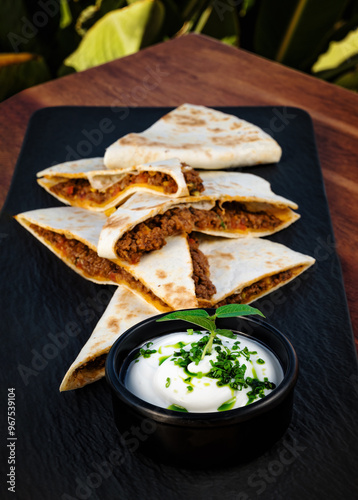 Close-up shot of gourmet Mexican-inspired dish featuring black plate with perfectly arranged golden-brown quesadilla slices. Beside plate, small black bowl holds creamy white dipping sauce 