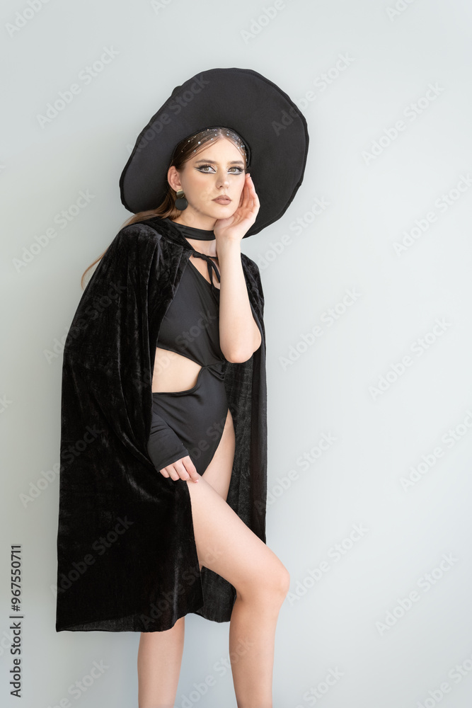 Young sexy woman in black bodysuit and cloak and witch hat. Dark makeup. White background. Halloween party, sexy witch Young sexy woman in black bodysuit and cloak and witch hat. Dark makeup. White background. Halloween party, sexy witch