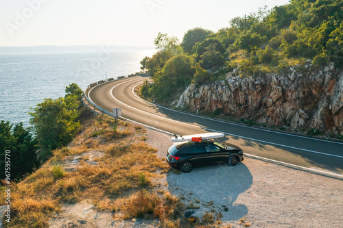 Black car with a paddleboard secured to its roof, parked at a scenic viewpoint on a high cliff