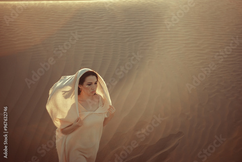 Ethereal woman in flowing white fabric stands amidst desert dunes at golden hour. Mysterious and alluring, her gaze fixed on the horizon. Soft light bathes the scene, creating a dreamlike atmosphere. 