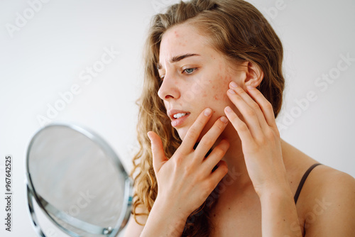 A young woman with curly hair is inspecting her face in a mirror, concentrating on blemishes and skin texture, showcasing a moment of self-care and concern. Skin problems. photo