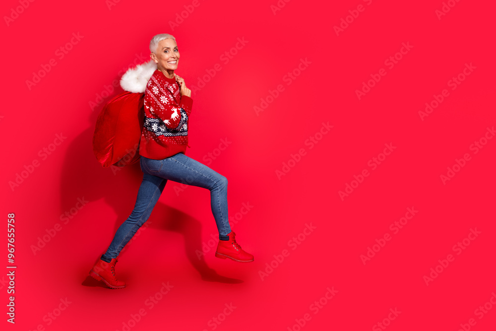 Full size photo of pretty retired female walking carry gift sack wear ornament sweater celebrate christmas isolated red color background Full size photo of pretty retired female walking carry gift sack wear ornament sweater celebrate christmas isolated red color background