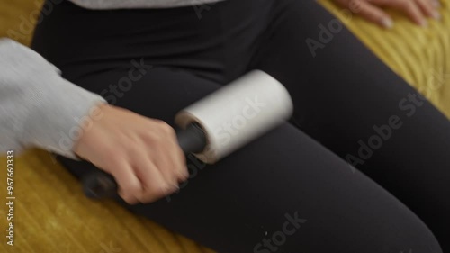 An adult woman uses a lint roller on her black pants while sitting on a yellow bed in a bedroom, showcasing the process of removing pet hair from clothes.