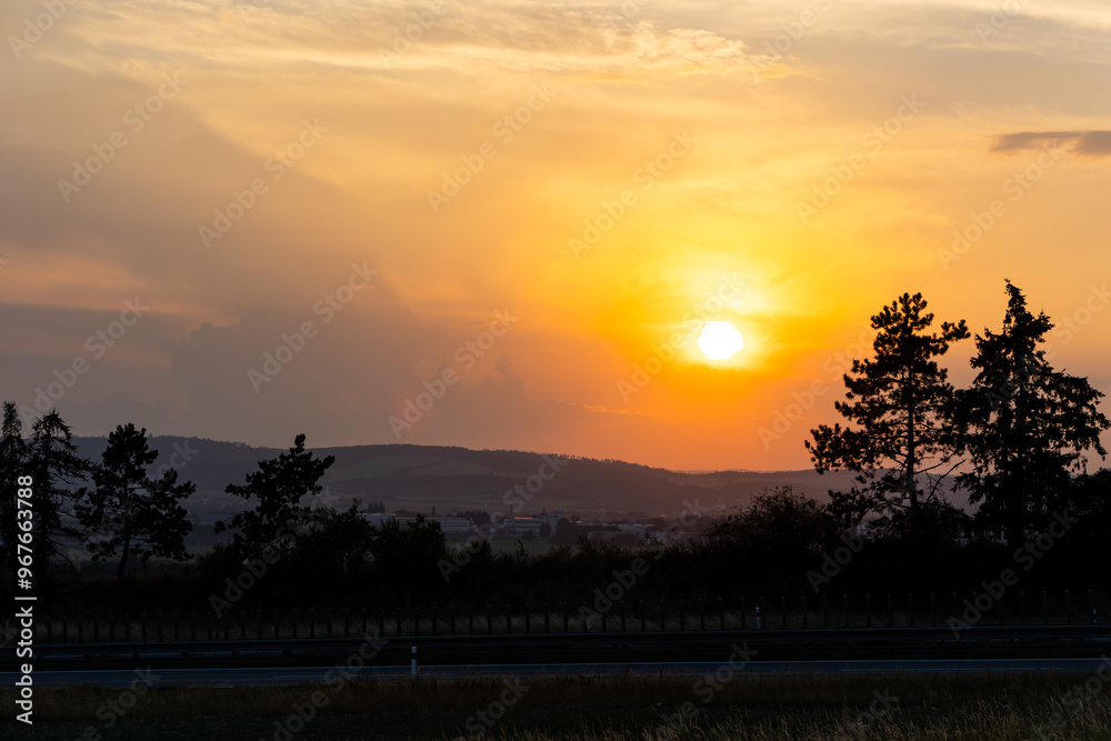 Orange sunset sun getting down amongs the trees over the road in the shade. Dramatic orange sky during the susnset.