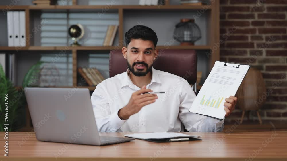 Serious indian office worker gesturing with hands while sitting at table with portable laptop. Earnest businessman pointing with pen on clickboard with paperwork. Serious indian office worker gesturing with hands while sitting at table with portable laptop. Earnest businessman pointing with pen on clickboard with paperwork.