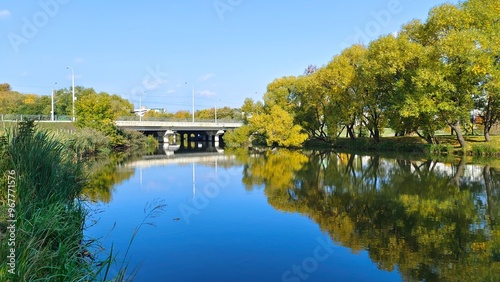 Willows and other deciduous trees grow on the grassy riverbank near the concrete bridge and are reflected in the water. The leaves on them have turned yellow. Reeds are growing in the water. Sunny 