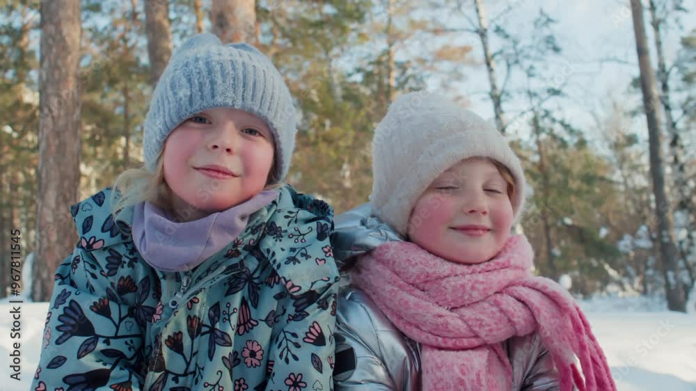 Chest up slowmo portrait of two adorable Caucasian girls in warm clothes posing in winter park on warm day and looking at camera Chest up slowmo portrait of two adorable Caucasian girls in warm clothes posing in winter park on warm day and looking at camera
