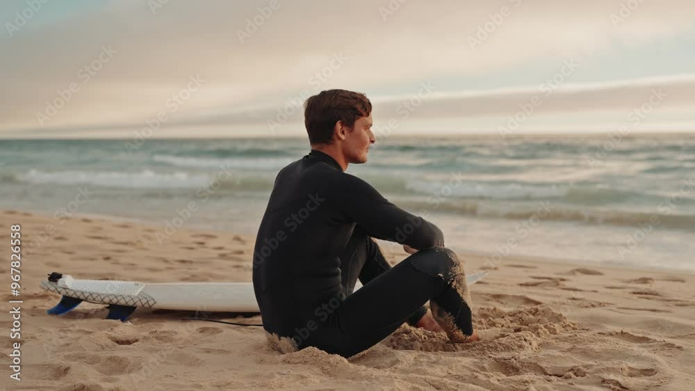 A surfer in a wetsuit sits on the beach with his surfboard, gazing out at the ocean waves and contemplating the next ride. A surfer in a wetsuit sits on the beach with his surfboard, gazing out at the ocean waves and contemplating the next ride.