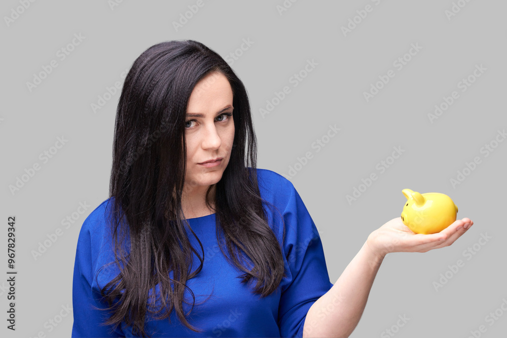 Woman in blue holding yellow piggy bank with skeptical expression against grey background