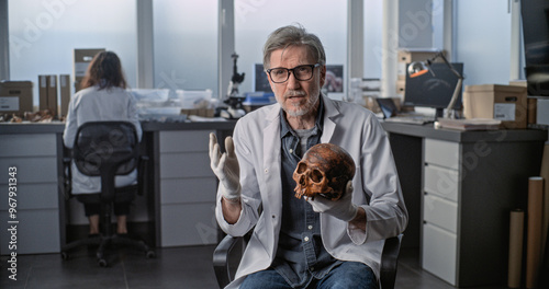 Tableau sur toile Mature scientist speaks to camera, shows fossil skull of ancient human during interview for popular science film in archaeological scientific lab