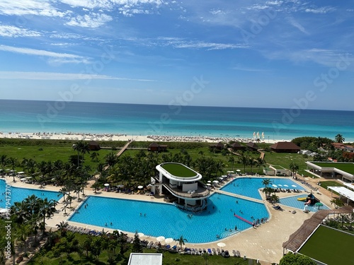 Aerial view of resort with pool, water slide, palm trees, and white sandy beach leading to turquoise ocean.