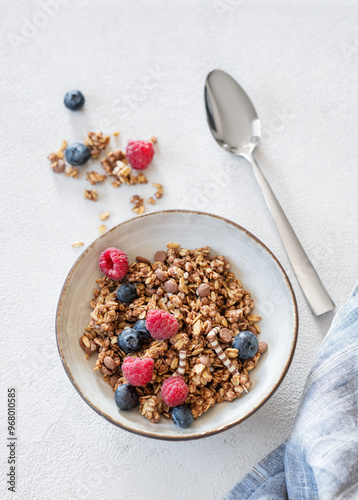 Canvas Print Dry muesli or granola, raspberries and blueberries in a bowl on a light background with fresh berries, spoon and morning sunlight