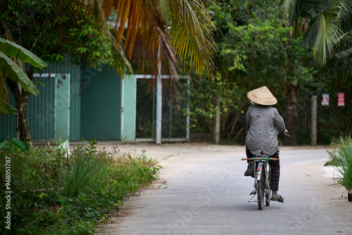 senior riding a bicycle through a Vietnamese village wearing traditional clothing and a Non la hat.